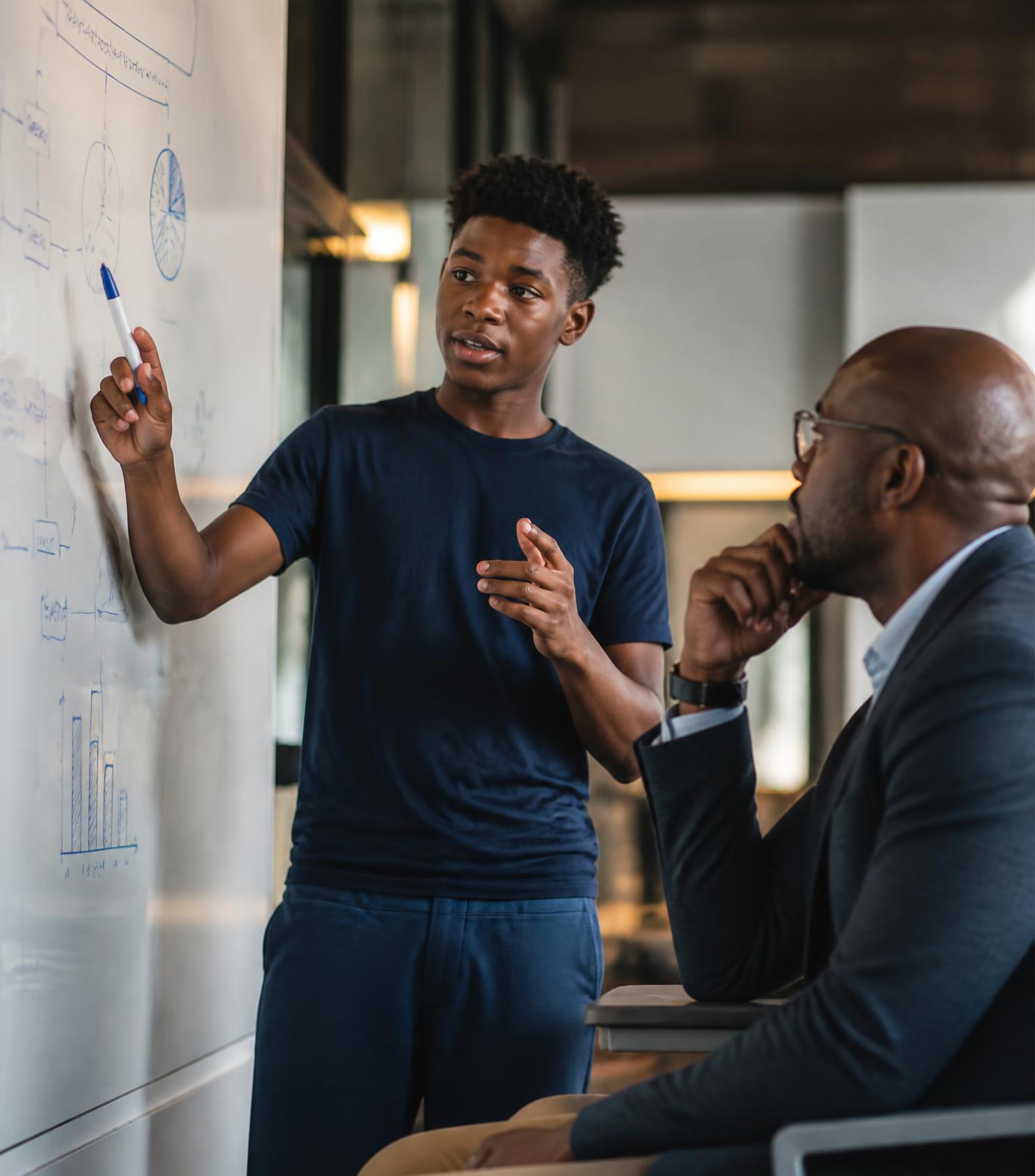 A student presenting a business plan on a whiteboard to an attentive adult mentor